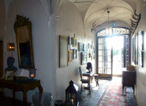 En gång i en gammal herrgård, tavlor hänger på väggen och en röd matta ligger på golvet. Gången leder till en öppen glasdörr som solen lyser igenom.